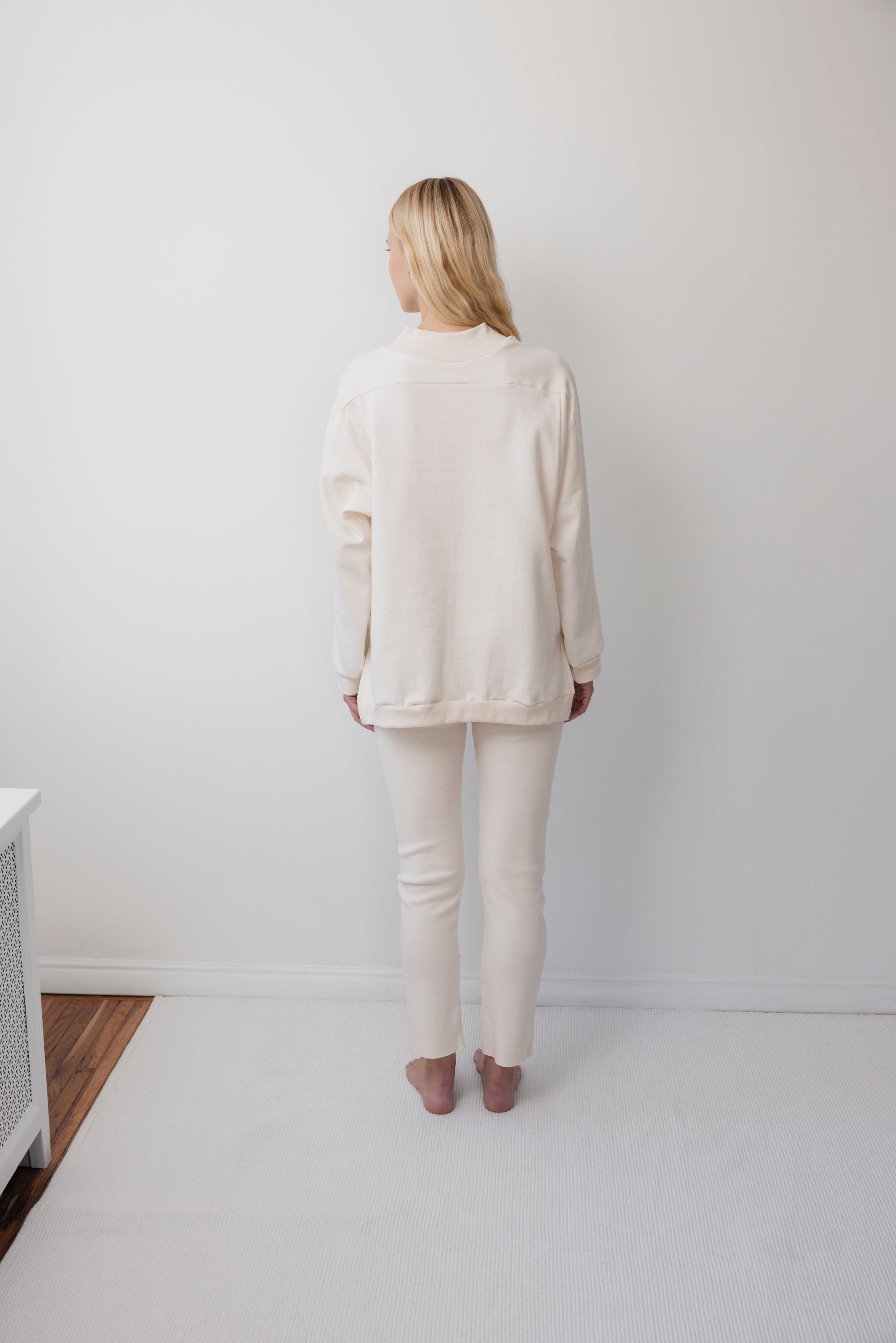A person with long blonde hair stands barefoot facing a white wall in a minimalist, bright room, wearing the oversized Big Sweatshirt : Natural by Wol Hide with matching pants featuring a wide rib hem.