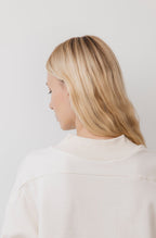 A woman with long, wavy blonde hair stands against a white background, wearing the Wol Hide Big Sweatshirt: Natural. The off-white oversized mock neck features a wide rib hem. She is shown from behind, with her profile turned slightly left.