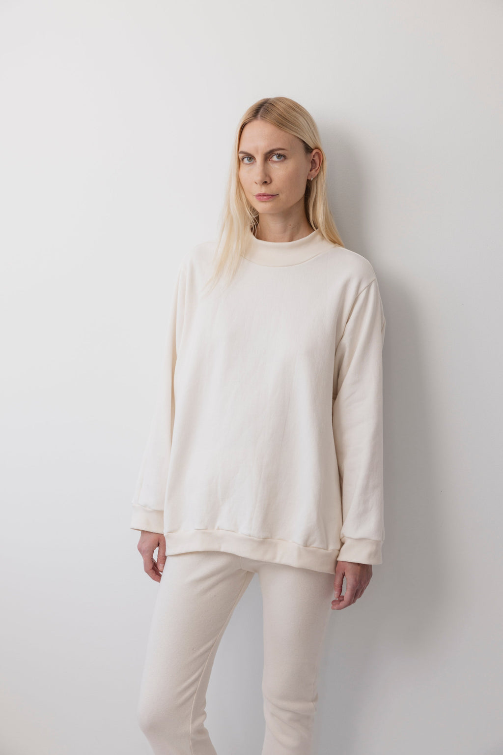 A person with long blonde hair stands against a plain white wall, wearing the Wol Hide Big Sweatshirt : Natural with a shoulder gusset seam and matching pants, looking directly at the camera with a neutral expression.