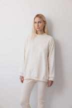 A person with long blonde hair stands against a plain white wall, wearing the Wol Hide Big Sweatshirt : Natural with a shoulder gusset seam and matching pants, looking directly at the camera with a neutral expression.