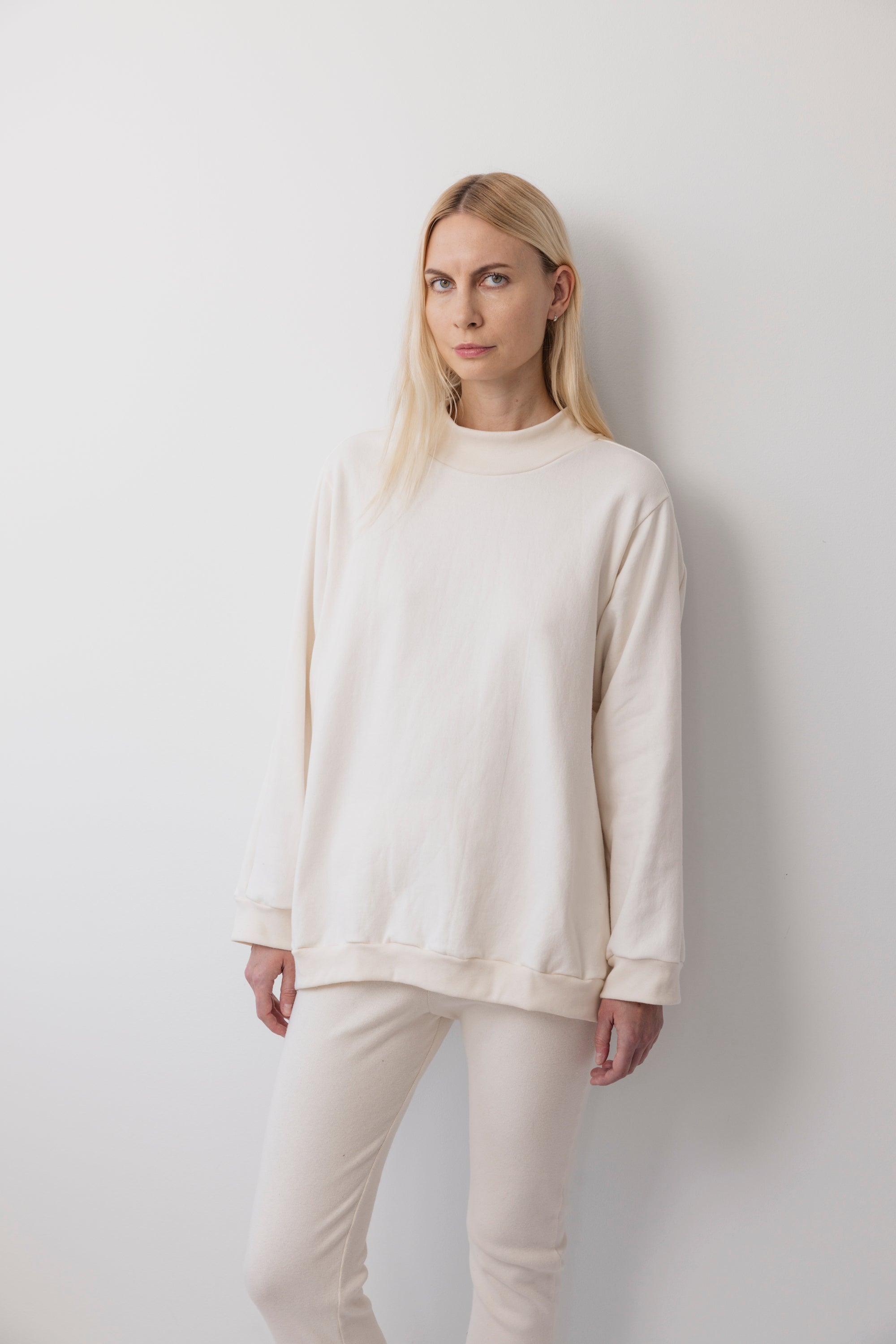 A person with long blonde hair stands against a plain white wall, wearing the Wol Hide Big Sweatshirt : Natural with a shoulder gusset seam and matching pants, looking directly at the camera with a neutral expression.