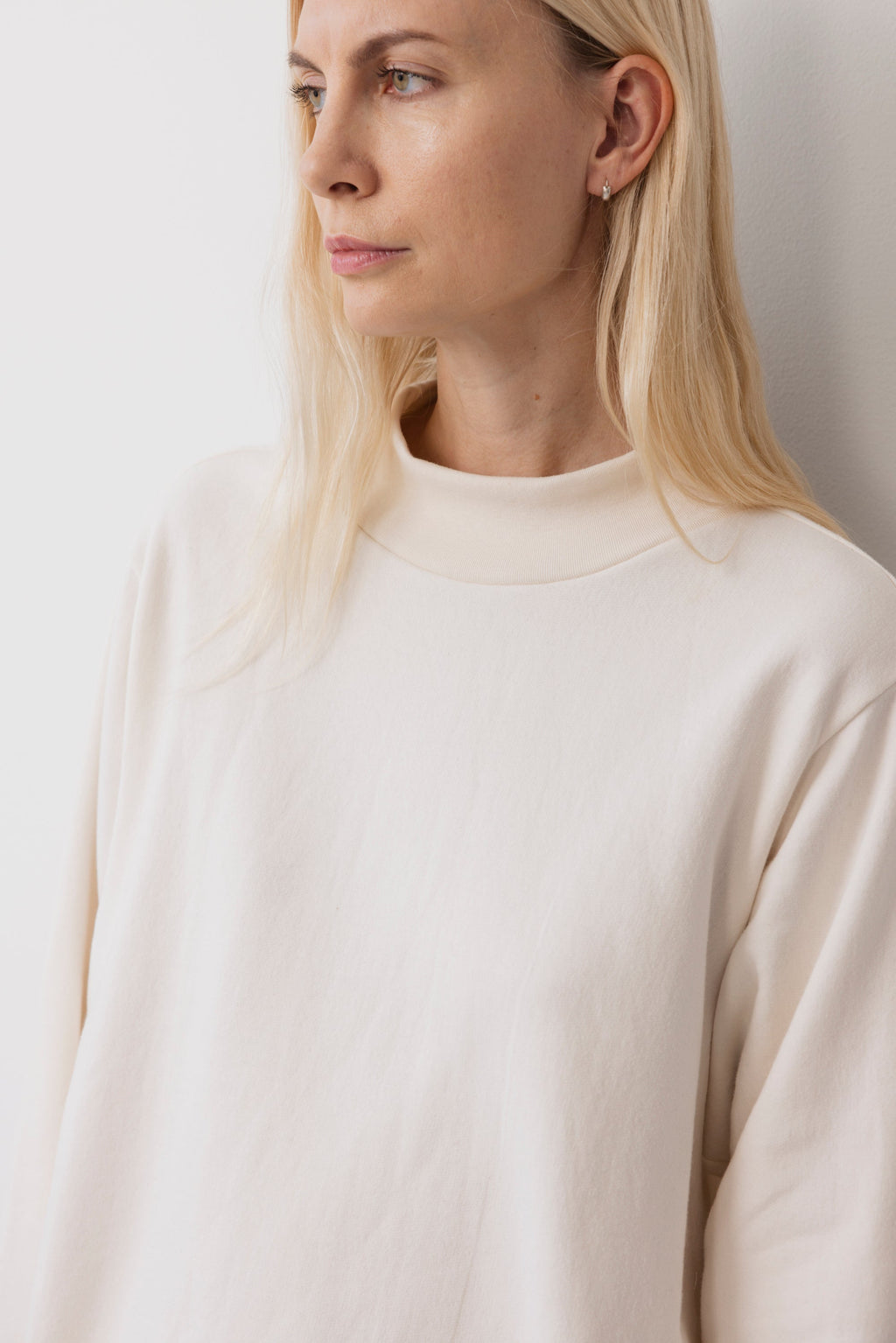 A person with long blonde hair models the Wol Hide Big Sweatshirt : Natural, featuring a wide rib hem and mock neck, while standing against a plain white background and looking slightly to the side.