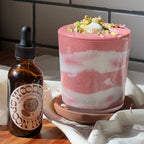 A glass with a pink and white smoothie, topped with whipped cream and chopped nuts, sits beside a WOODEN SPOON HERBS Rose-Colored Glasses dropper bottle on a sunlit wooden surface.