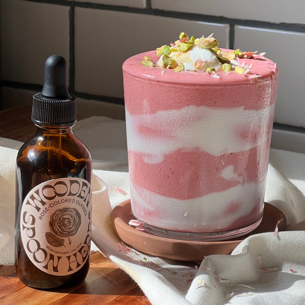A glass with a pink and white smoothie, topped with whipped cream and chopped nuts, sits beside a WOODEN SPOON HERBS Rose-Colored Glasses dropper bottle on a sunlit wooden surface.