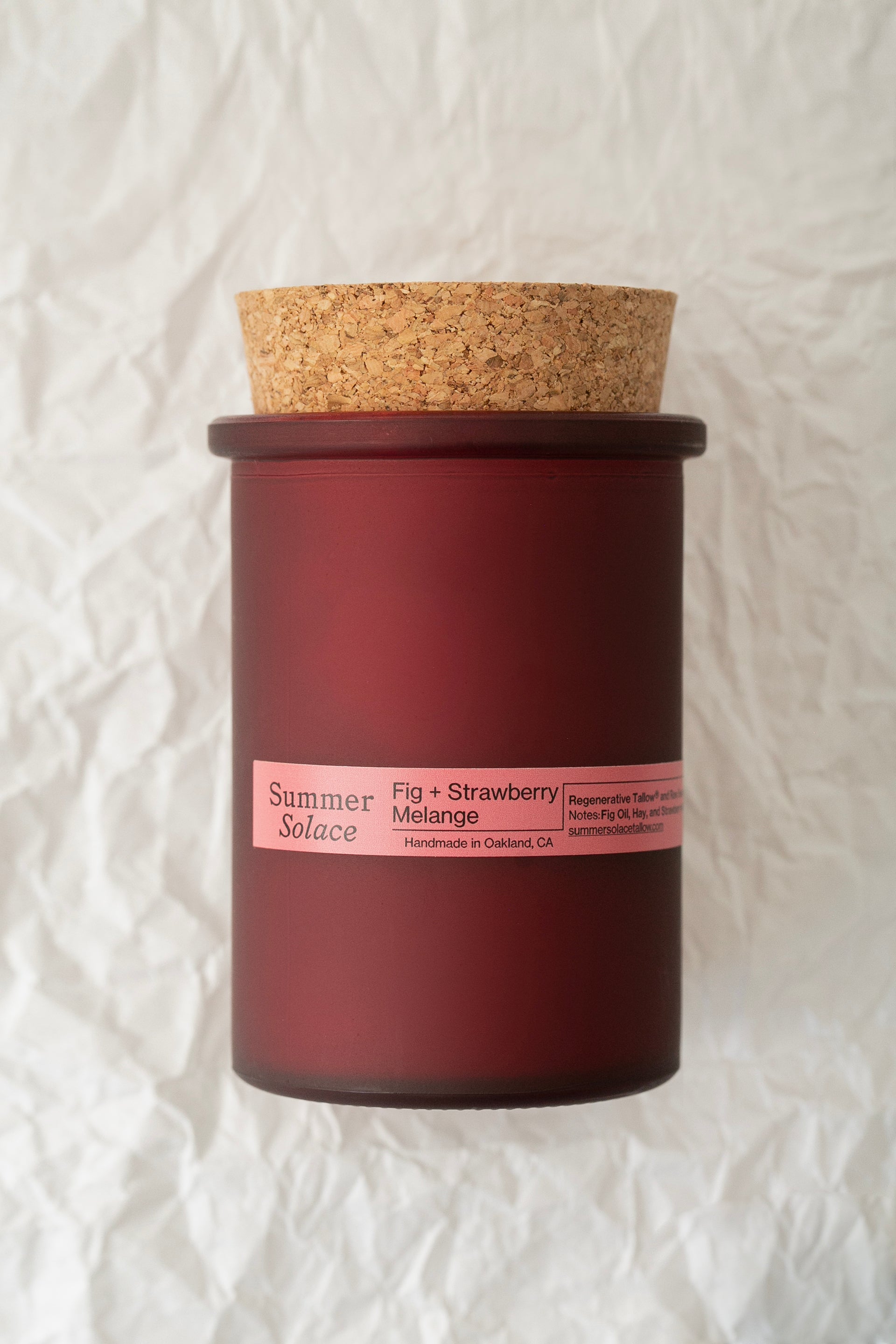 A red, frosted glass container with a cork lid bearing the labels "Summer Solace Tallow" and "Fig and Strawberry Melange Candle - Regenerative Tallow® 6 oz." is set against crumpled white paper. This slow-burning candle, handmade in Oakland, CA, enchants with its fruity fragrance.