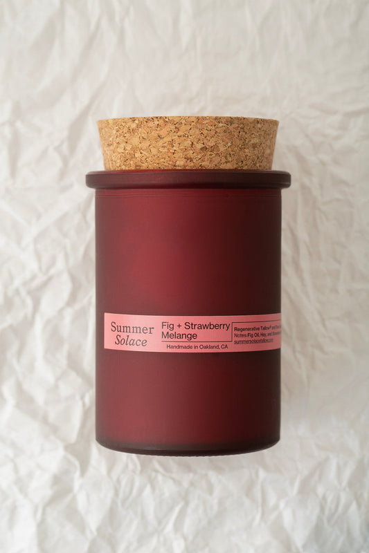 A red, frosted glass container with a cork lid bearing the labels "Summer Solace Tallow" and "Fig and Strawberry Melange Candle - Regenerative Tallow® 6 oz." is set against crumpled white paper. This slow-burning candle, handmade in Oakland, CA, enchants with its fruity fragrance.