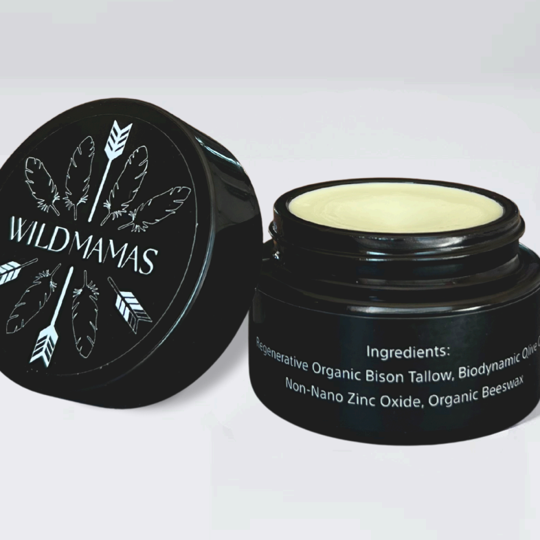 A black jar of Wild Mamas Sunscreen—Wild Bison Tallow Balm sits open, unveiling the light-yellow balm inside. Its label highlights wild bison tallow and non-nano zinc oxide for a regenerative, organic sunscreen with natural ingredients.