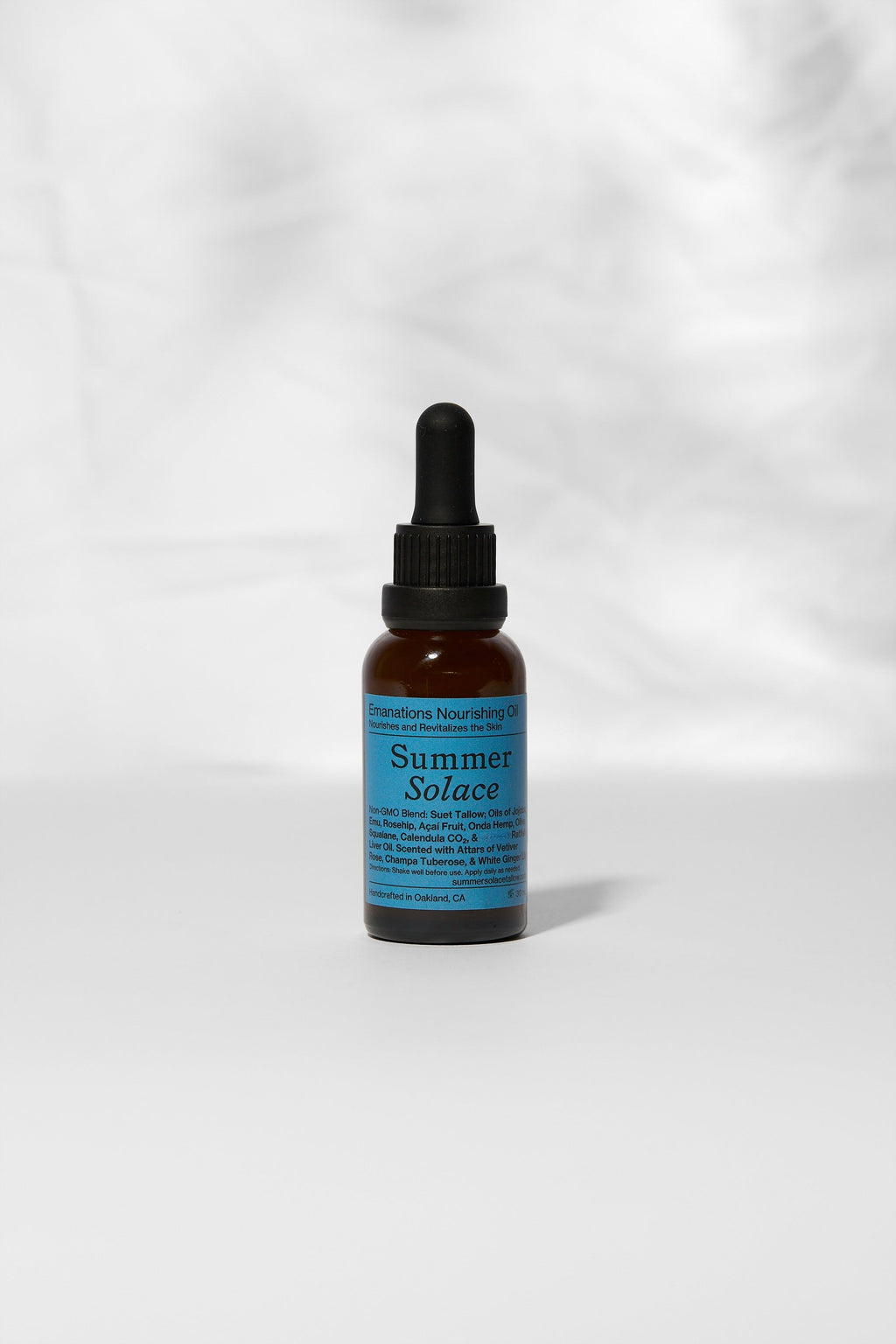 A small amber glass dropper bottle with a black cap and a blue label reading "Summer Solace Tallow" stands upright on a white surface, holding Emanations Profound Face Oil—deeply nourishing tallow, emu oil, champa & white ginger lily.