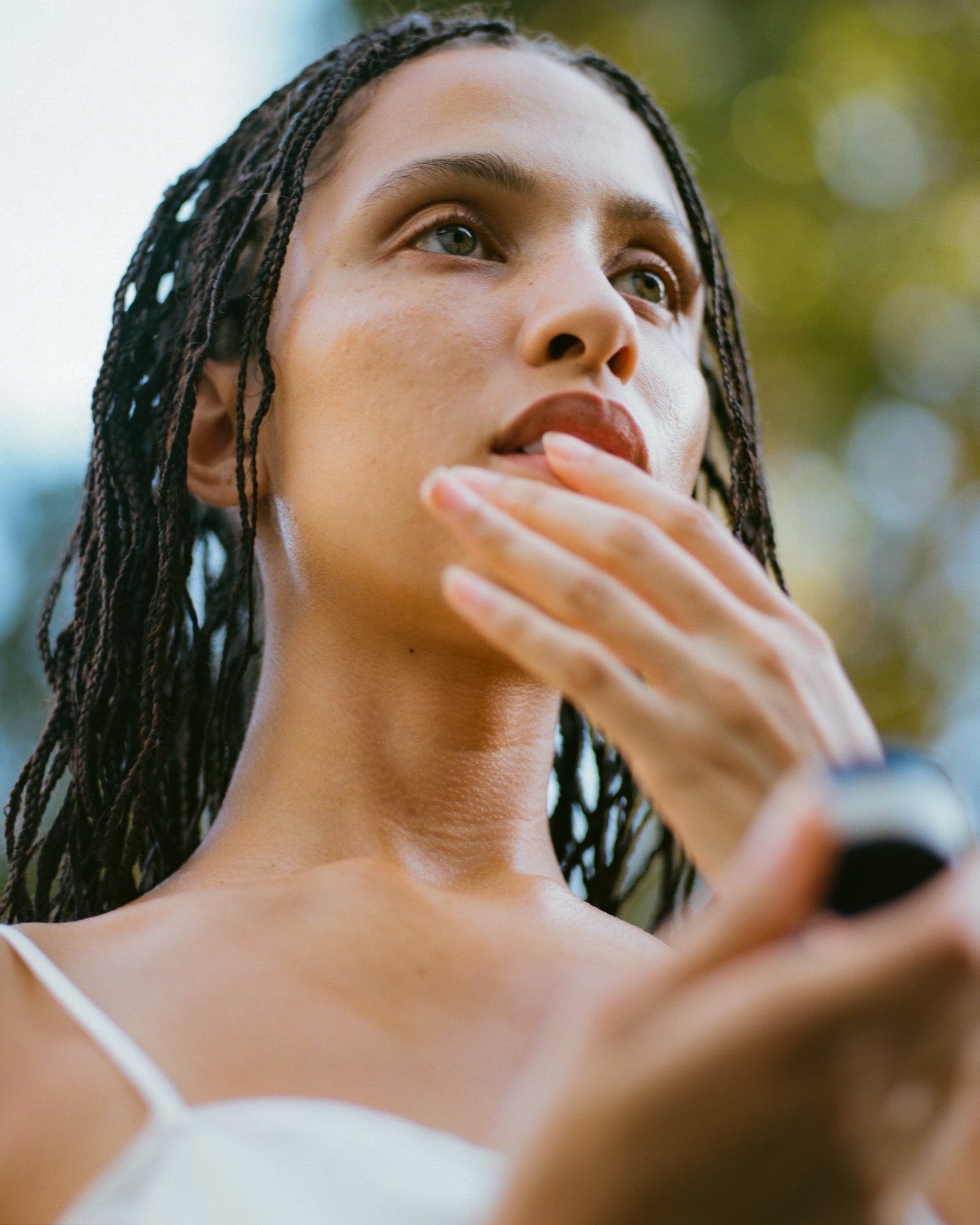 A person with braided hair applies Surya's Calming Lip Therapy outdoors, wearing a white top. The blurred green-yellow background suggests nature, and the photo is shot from a low angle.