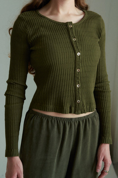 A woman indoors wears the LA RELAXED Pointelle Cardigan, an olive green ribbed long-sleeve top with pearlescent buttons, paired with matching loose pants. The cropped image shows her from below the shoulders without showing her face.
