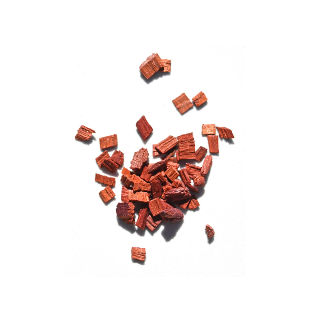 Reddish-brown organic sandalwood chips, key to Daughter Alchemy’s Calm Ground® Botanical Calming Oil, are scattered on a white background.