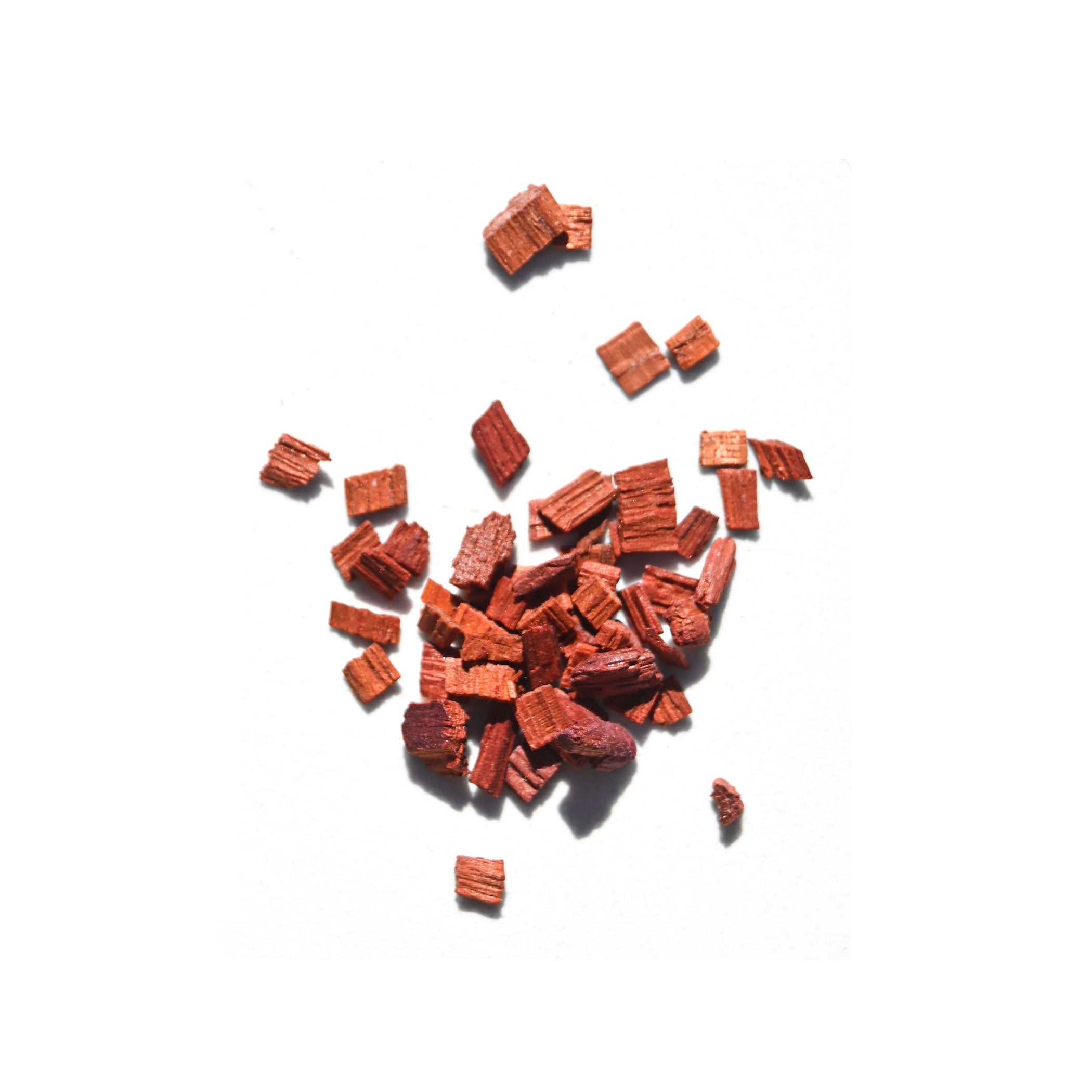 Reddish-brown organic sandalwood chips, key to Daughter Alchemy’s Calm Ground® Botanical Calming Oil, are scattered on a white background.