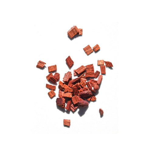 Reddish-brown organic sandalwood chips, key to Daughter Alchemy’s Calm Ground® Botanical Calming Oil, are scattered on a white background.