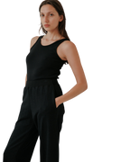 A woman with long brown hair wears the Wol Hide Scoop Back Rib Tank in black with matching pants, standing with one hand in her pocket against a light background, highlighting the effortless style of sustainable fashion.