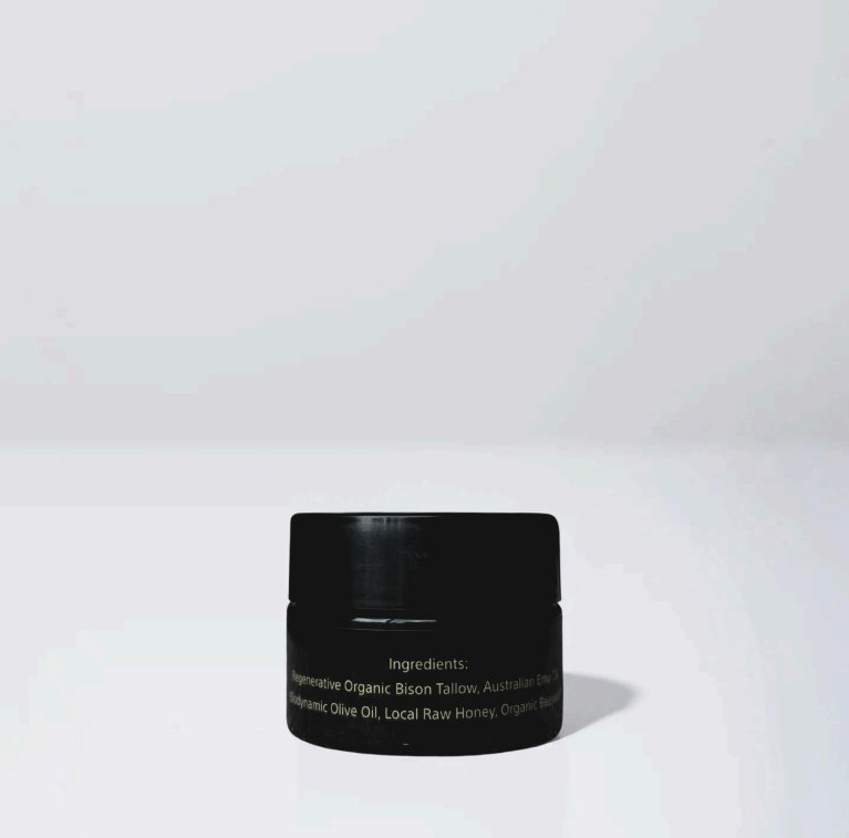 A small black jar of Wild Mamas Honey Bee (Raw Honey) - Wild Bison Tallow Balm with a faint label sits on a white surface against a light gray background. The text on the jar is hard to read due to low lighting and reflections.
