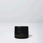 A small black jar of Wild Mamas Honey Bee (Raw Honey) - Wild Bison Tallow Balm with a faint label sits on a white surface against a light gray background. The text on the jar is hard to read due to low lighting and reflections.