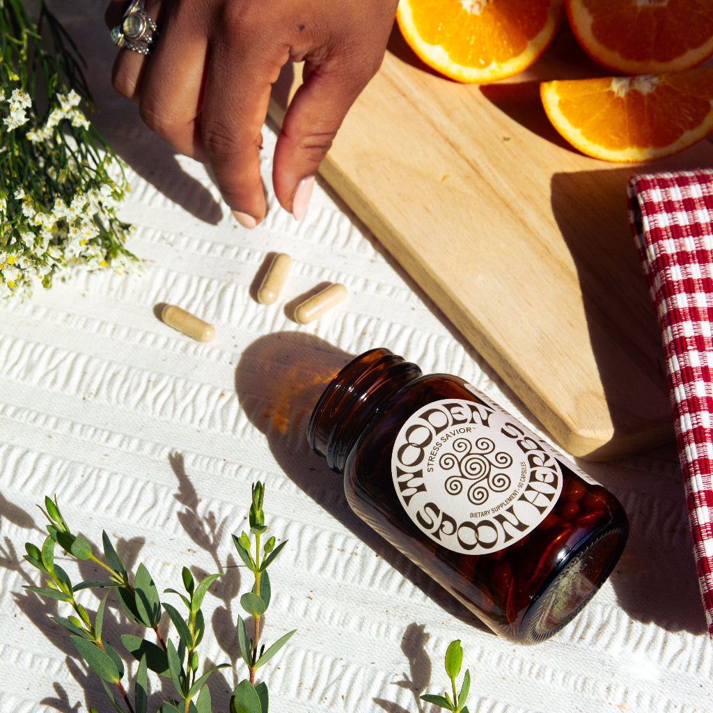 A hand reaches for Stress Savior capsules spilled from a WOODEN SPOON HERBS bottle on a textured white surface, surrounded by sliced oranges, flowers, and greenery—evoking stress relief through adaptogenic herbs.