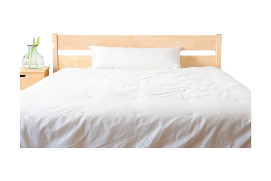A bed with a light wood headboard features a white pillow and the Shepherd's Dream Summer Weight Wool Comforter. To the left, a wooden nightstand holds a glass vase filled with green leaves.