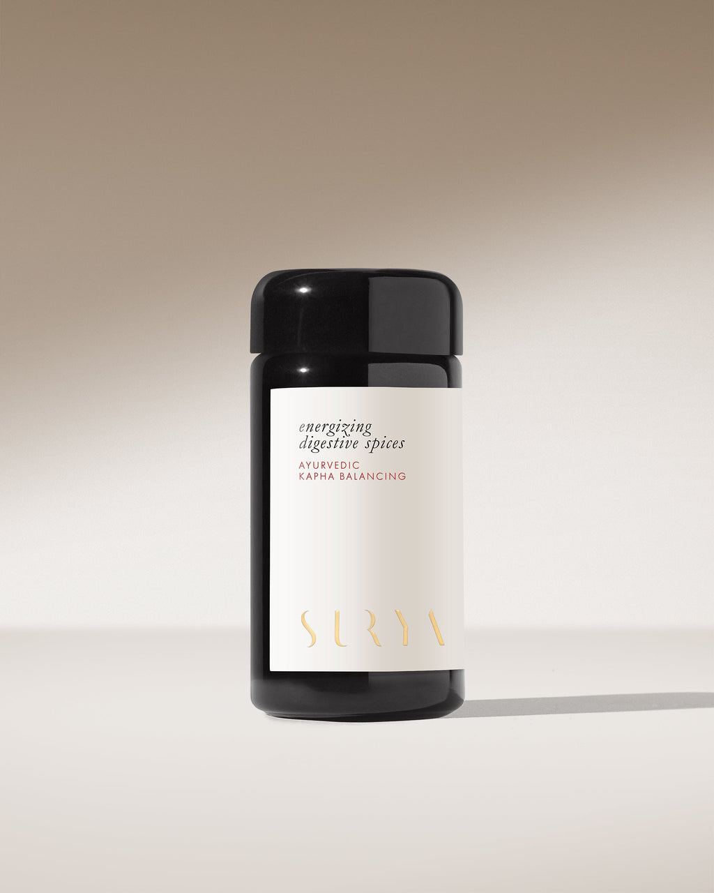 A dark glass jar with a white label reading "Energizing Digestive Spices" and "Surya" in gold sits against a neutral, softly lit background—ideal as a Kapha-balancing Ayurvedic spice blend or savory condiment for your cooking.