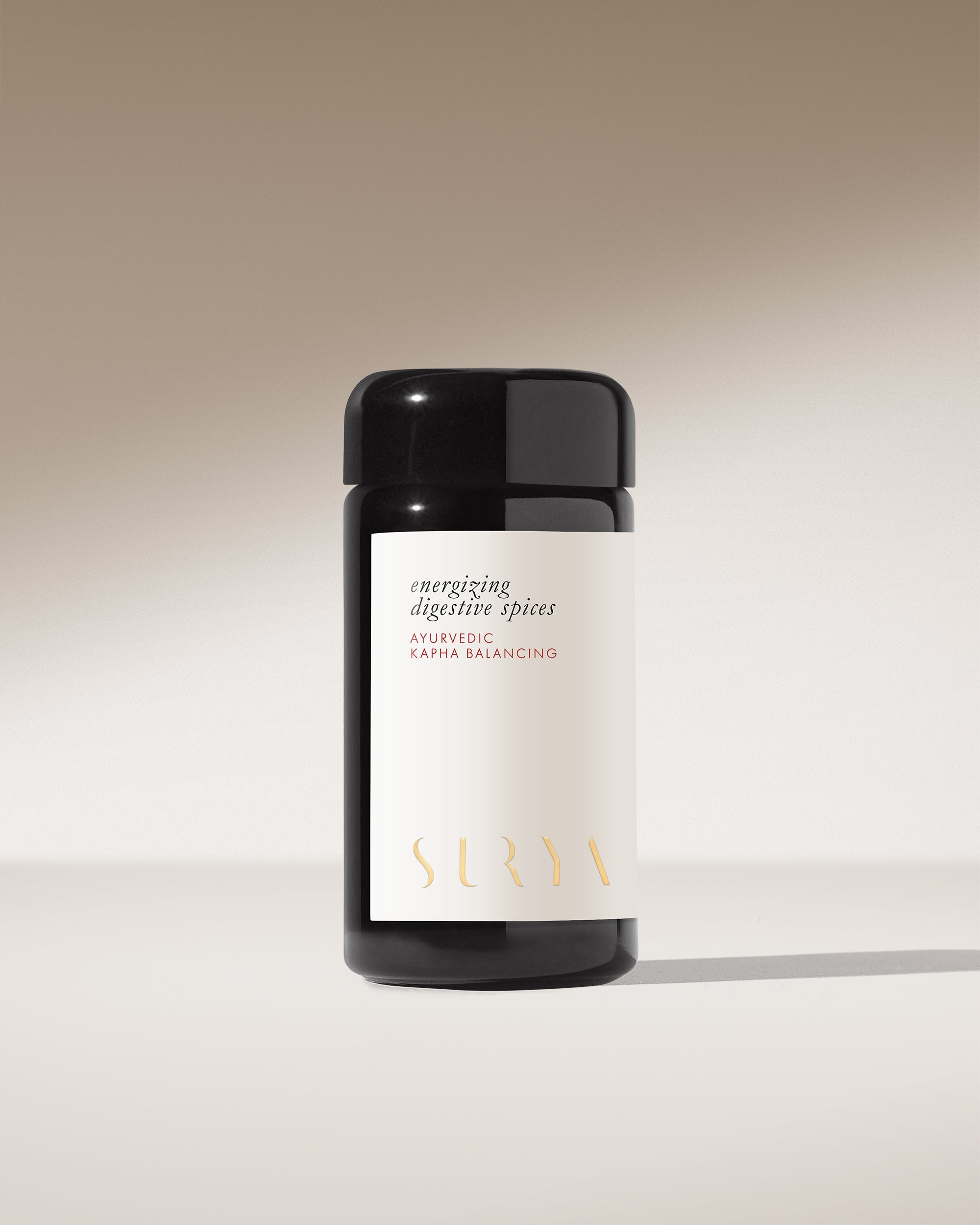 A dark glass jar with a white label reading "Energizing Digestive Spices" and "Surya" in gold sits against a neutral, softly lit background—ideal as a Kapha-balancing Ayurvedic spice blend or savory condiment for your cooking.