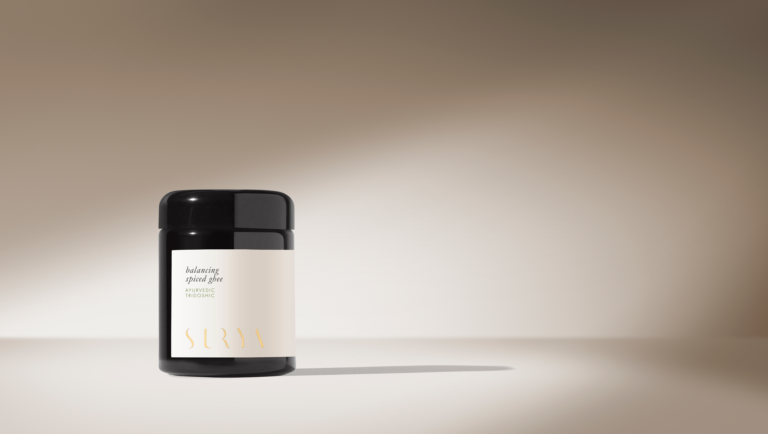 A dark jar with a white label reading "Spiced Ghee" and "Surya" sits on a smooth surface, evoking the purity of organic ghee used in Ayurvedic cooking, against a neutral beige gradient with soft shadows.