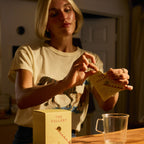 A woman with short blonde hair opens a yellow packet of The Fullest Saffron Latte 10 Single-Serving Sachets above a clear glass mug on a wooden countertop, warmly lit indoors.