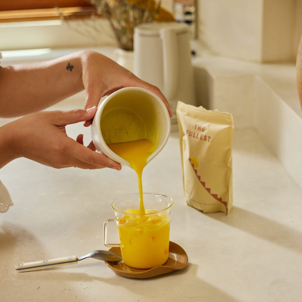A person pours The Fullest Saffron Latte 30-Day Supply, a caffeine-free bright yellow drink, from a white pitcher into a glass mug on a wooden plate, with a spoon and the beige “The Fullest” packet in the background for mood support.