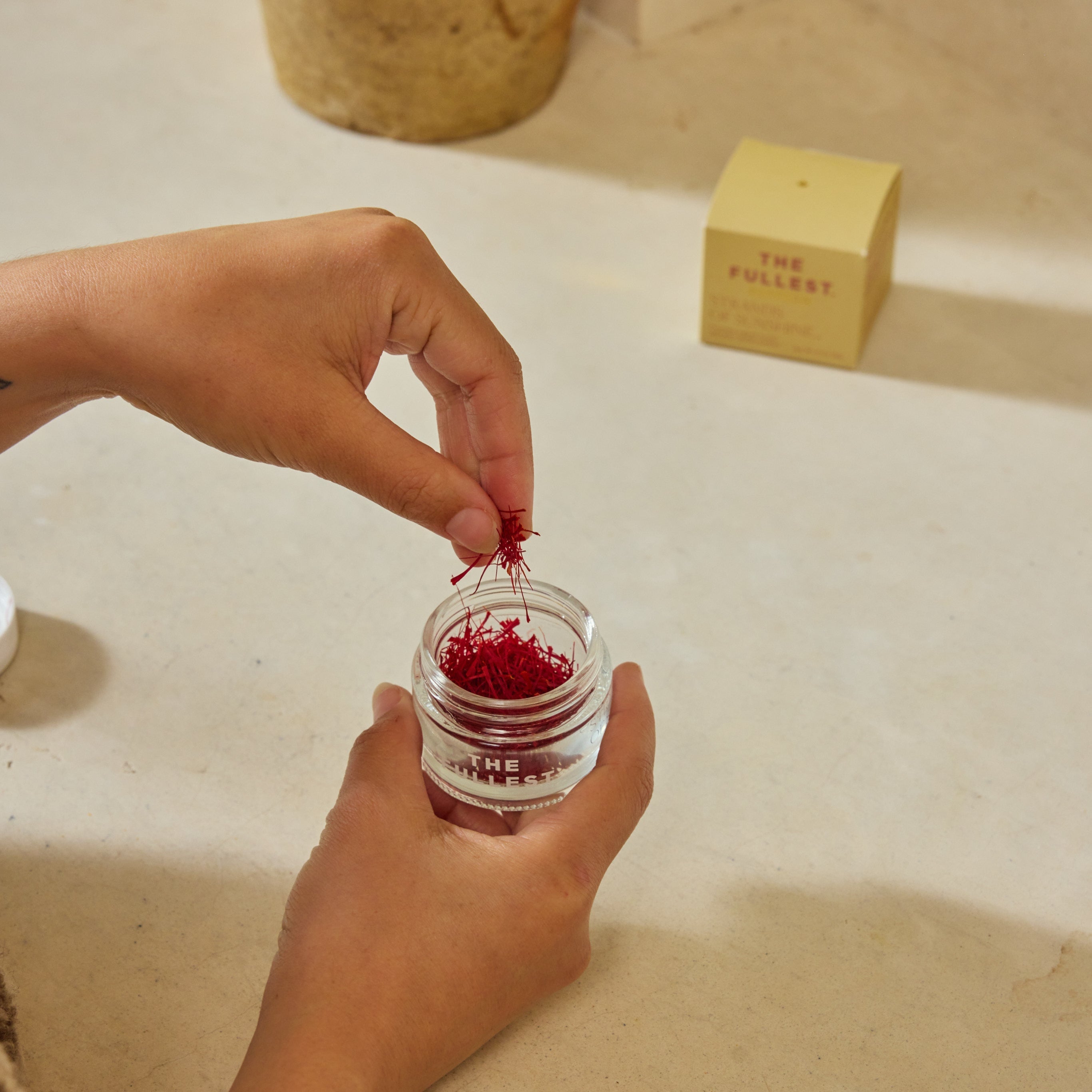 A person takes saffron threads from a small glass jar of Saffron Threads by The Fullest, with the brand’s beige box visible on a light surface in the background.