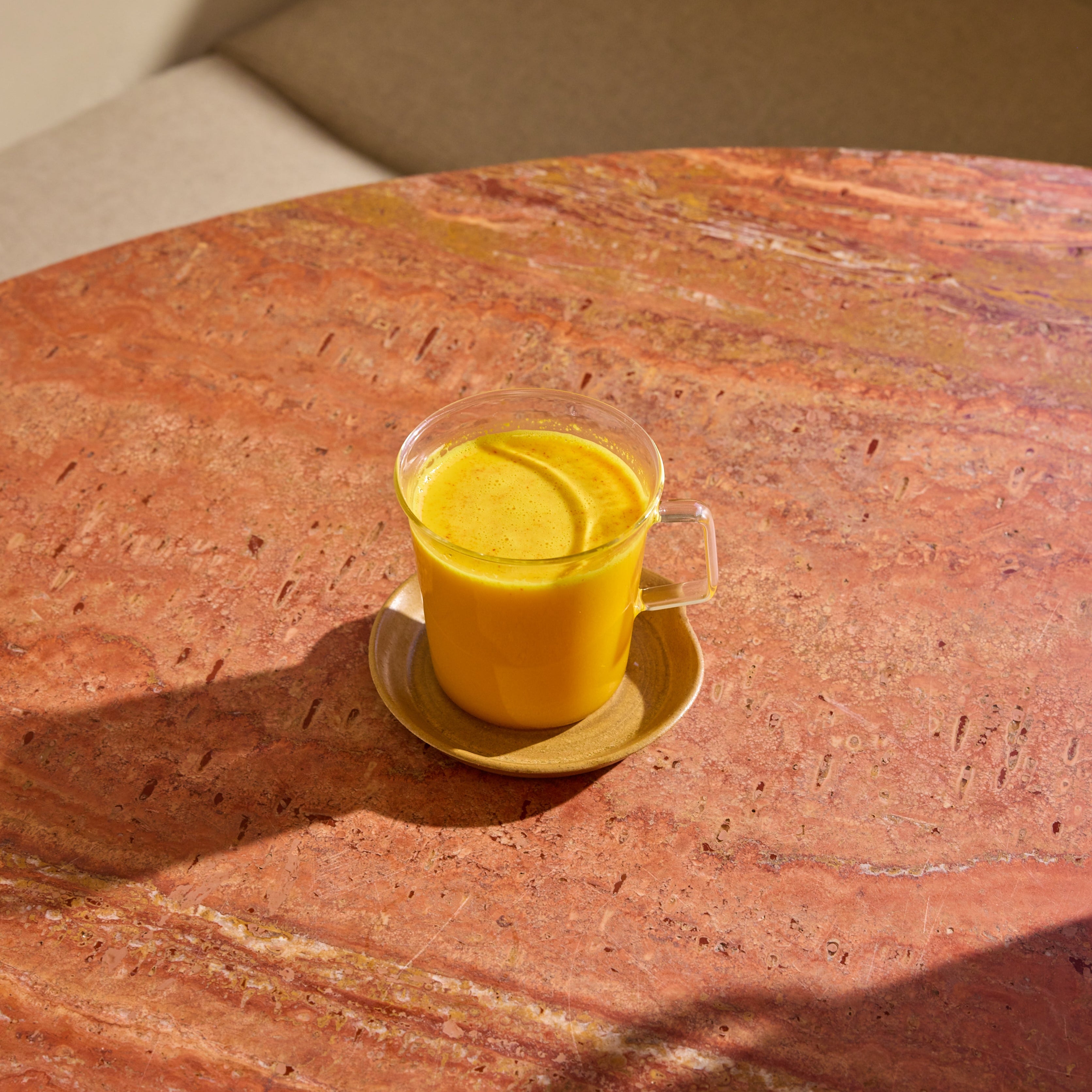 A glass mug of The Fullest's caffeine-free Saffron Latte 30-Day Supply sits on a round, pinkish-brown marble table in bright sunlight, served on a matching saucer.