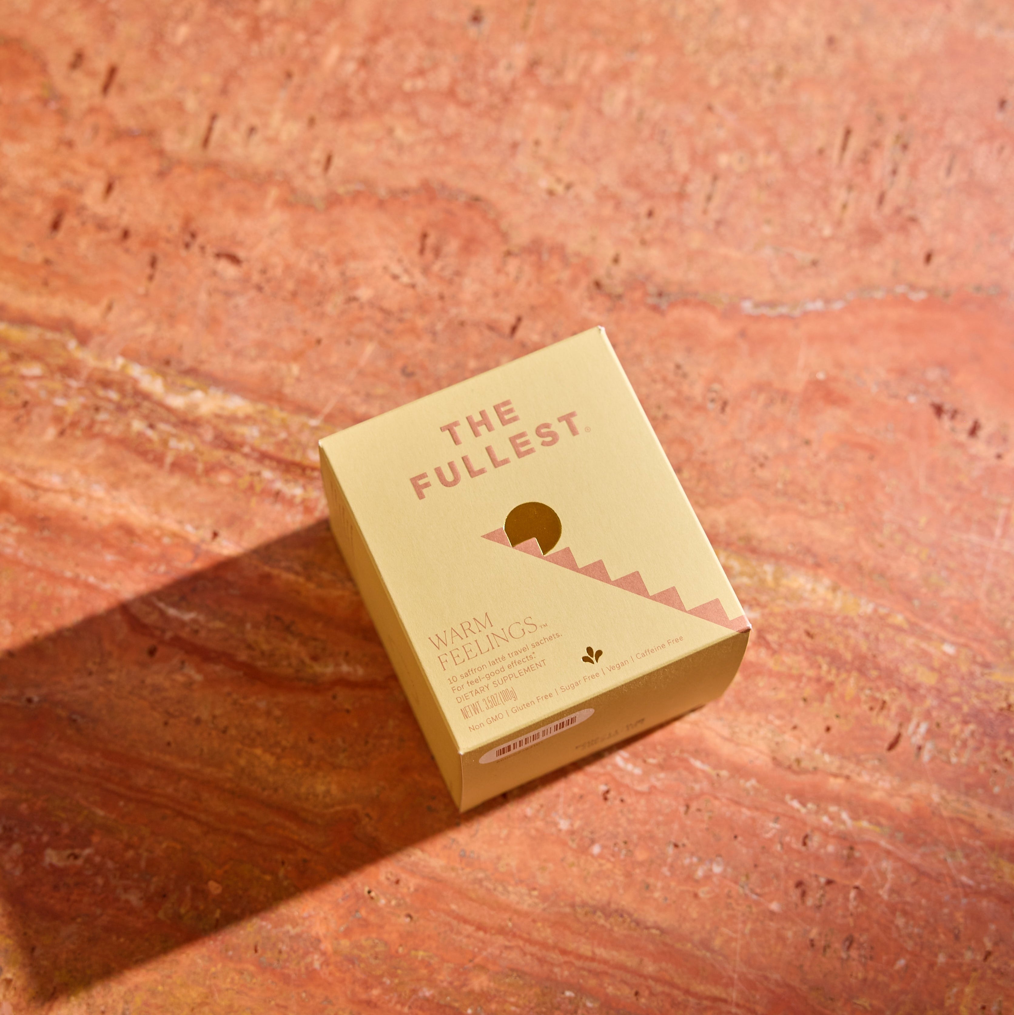 A small beige box labeled "The Fullest" sits on marbled orange, featuring pink geometric designs and reading "Warm Feelings™ Saffron Latte 10 Single-Serving Sachets," a caffeine-free blend for gentle mood support.