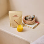 A packet of The Fullest Saffron Latte 30-Day Supply sits next to a small glass of caffeine-free saffron latte, a sage bundle in a dish, and two closed books on a light surface against a beige wall.