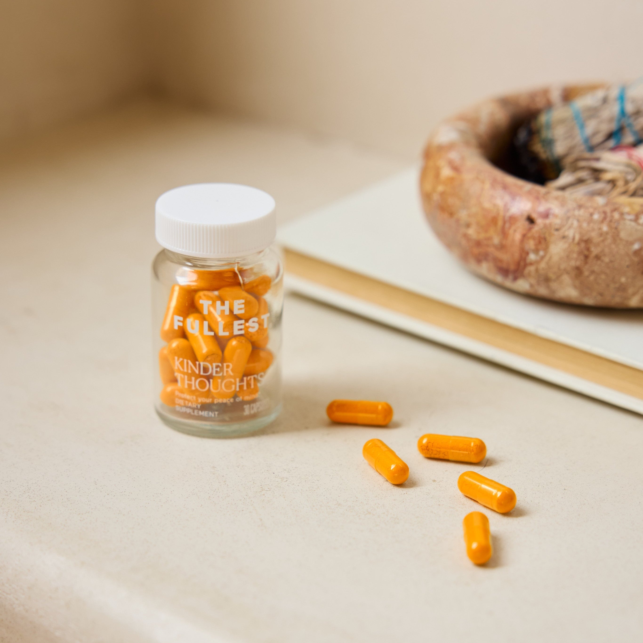 A clear bottle of The Fullest Saffron Capsules for mood support sits on a beige surface, with several capsules scattered nearby next to a round decorative bowl.