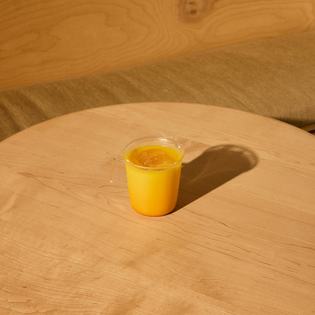 A clear cup of The Fullest Saffron Latte sits on a light wooden table beside a cushioned bench and wooden wall, its vibrant yellow-orange color highlighted by sunlight. This caffeine-free drink comes in 10 single-serving sachets.