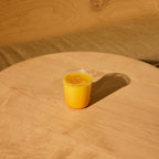 A clear cup of The Fullest Saffron Latte sits on a light wooden table beside a cushioned bench and wooden wall, its vibrant yellow-orange color highlighted by sunlight. This caffeine-free drink comes in 10 single-serving sachets.