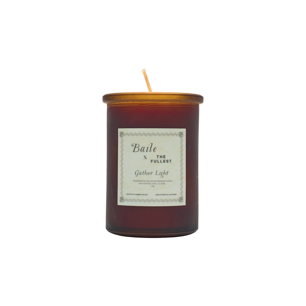 A brown glass jar candle called "Gather Light - Saffron Beeswax Tallow Candle" by Baile features a gold lid and white label. The unlit, centered wick is non-toxic, and the product is shown against a plain white background.