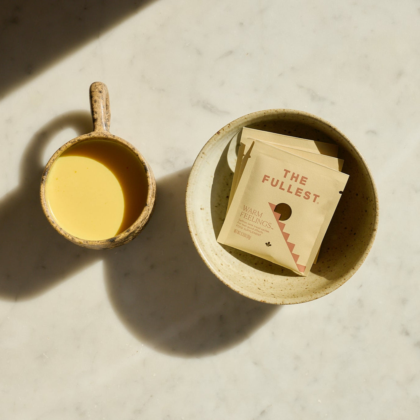 A beige ceramic cup of creamy yellow Saffron Latte from The Fullest sits beside a speckled bowl with two yellow Saffron Latte 10 Single-Serving Sachets, all on a light marble surface illuminated by gentle sunlight.