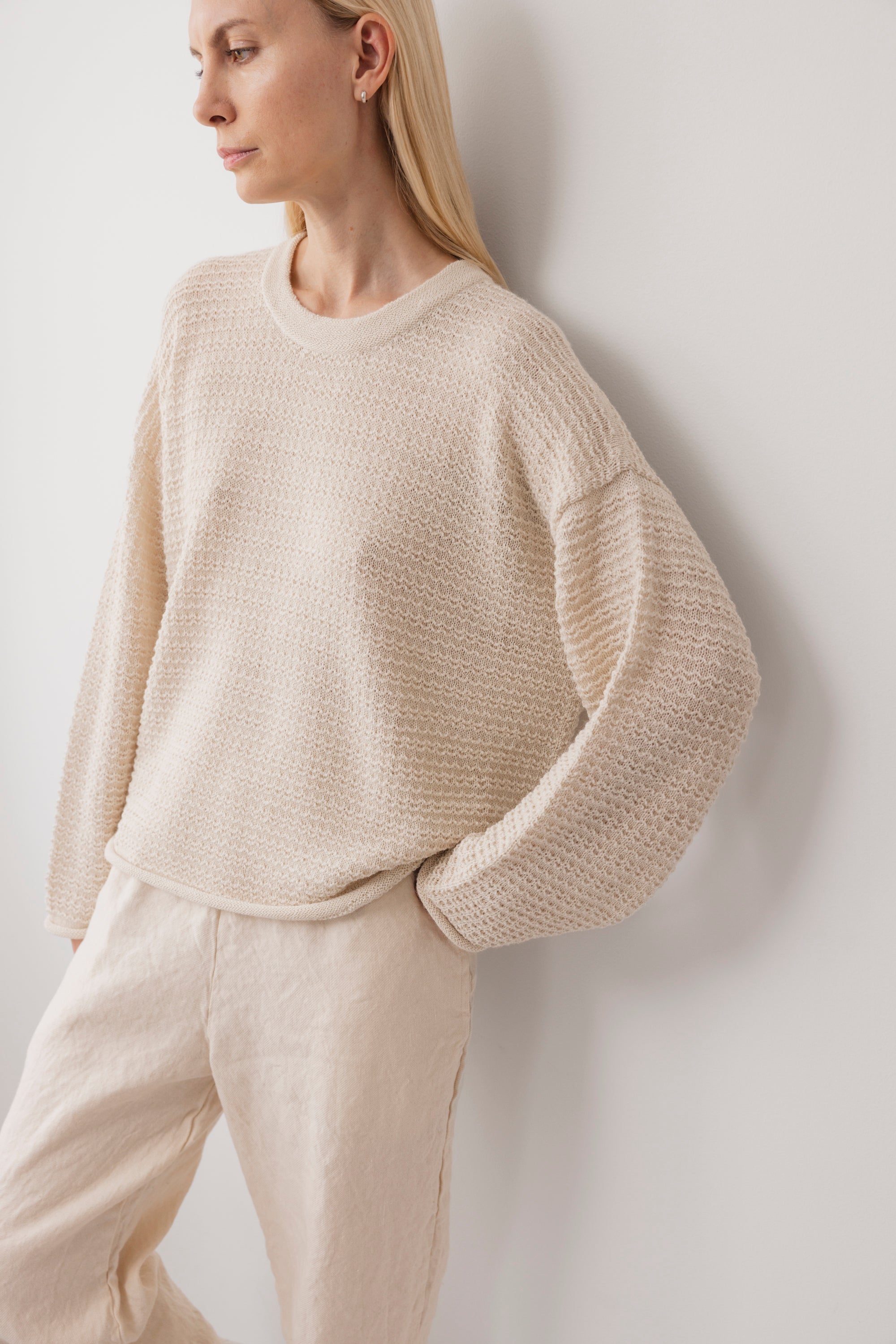A person wears the Wol Hide Thermal Sweater in Natural and light beige pants, standing against a white wall with hands in pockets. The look is relaxed and minimalist.