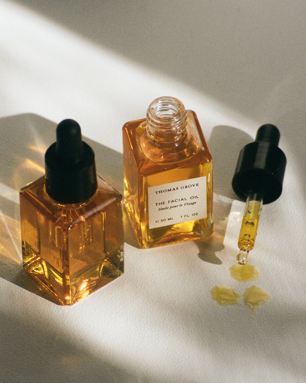 Two glass dropper bottles of THOMAS GROVE SKIN THE FACIAL OIL sit on a light surface; one is open with the dropper nearby holding golden oil. The label highlights its premium organic skincare formula.