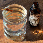 A dropper of liquid hovers above a clear glass of water beside an amber bottle labeled "WOODEN SPOON HERBS TRANQUILITY TONIC," a calming herbal blend resting on a wooden surface in sunlight.