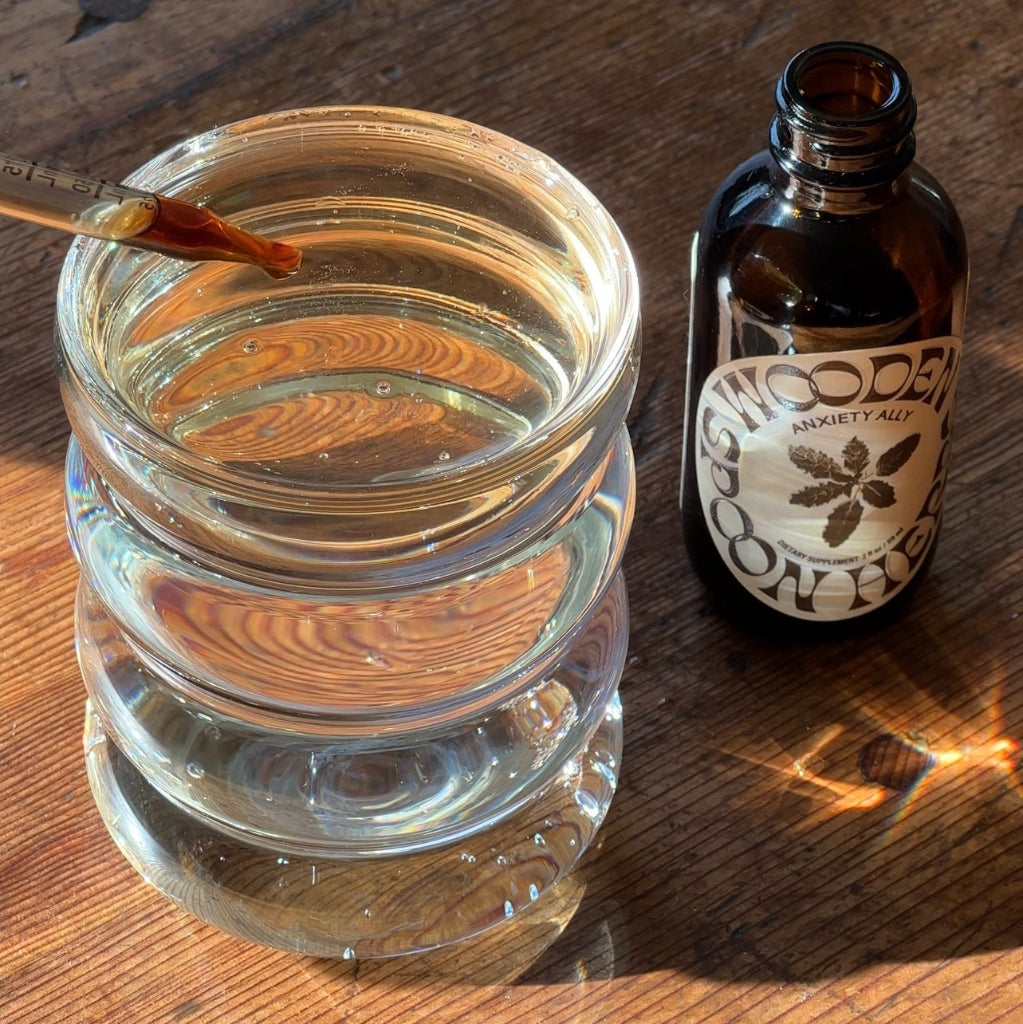 A dropper of liquid hovers above a clear glass of water beside an amber bottle labeled "WOODEN SPOON HERBS TRANQUILITY TONIC," a calming herbal blend resting on a wooden surface in sunlight.