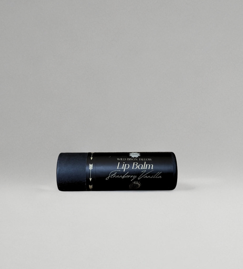 A black cylindrical tube of Wild Mamas Strawberry Vanilla Tallow Lip Balm rests horizontally on a light gray surface. The label features white and gold text, emphasizing its regenerative organic ingredients.