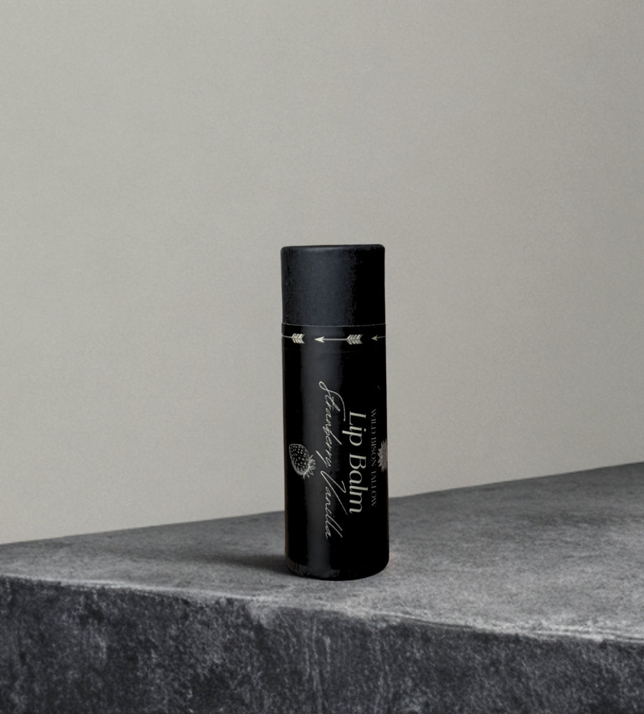 A black cylindrical tube of Wild Mamas Strawberry Vanilla Tallow Lip Balm with white text stands upright on a textured gray stone, set against a plain light gray background.