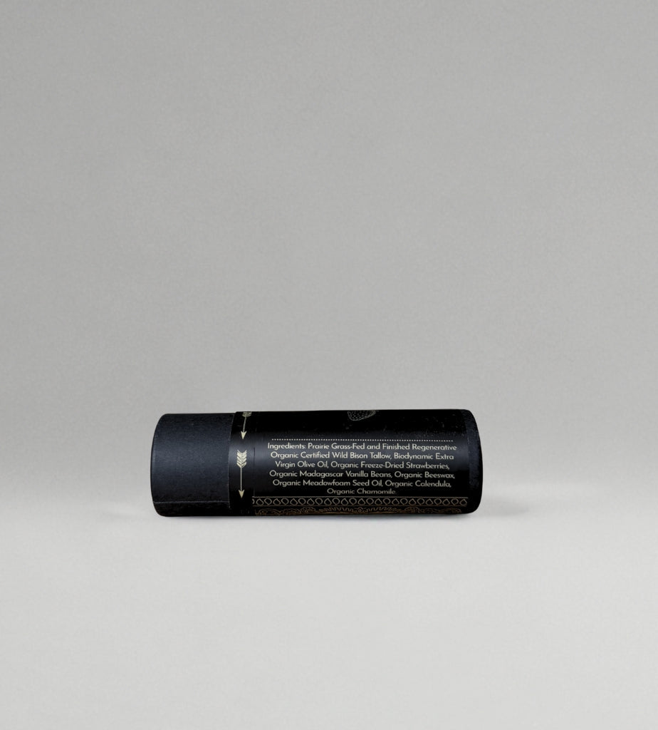 A black cylindrical container with gold accents from Wild Mamas lies horizontally on a light gray background. Its label, in small white letters, highlights the nourishing and regenerative organic benefits of Strawberry Vanilla Tallow Lip Balm.