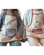 Two kids sit side by side wearing Baile x Casa Carini Baile Homegrown Aprons—one with "Homegrown" and a checkered pocket, the other with a house design and matching trim—both beautifully crafted in Italy.