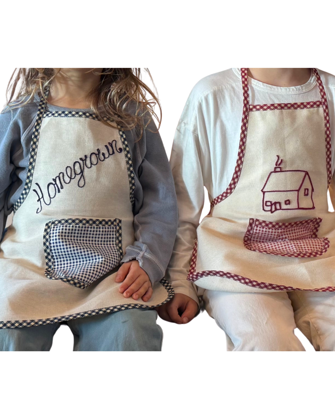 Two kids sit side by side wearing Baile x Casa Carini Baile Homegrown Aprons—one with "Homegrown" and a checkered pocket, the other with a house design and matching trim—both beautifully crafted in Italy.