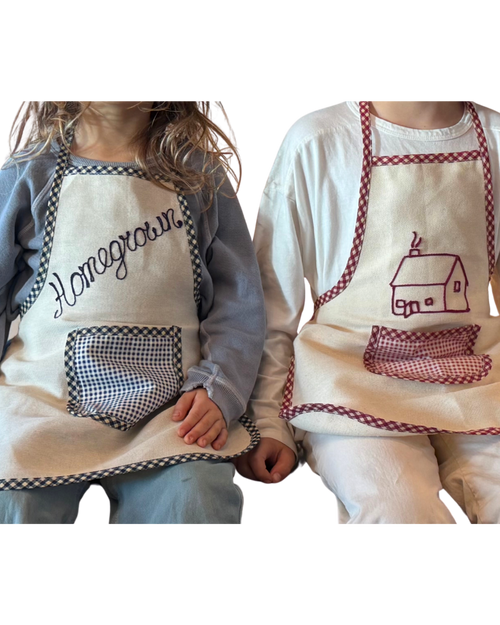 Two kids sit side by side wearing Baile x Casa Carini Baile Homegrown Aprons—one with "Homegrown" and a checkered pocket, the other with a house design and matching trim—both beautifully crafted in Italy.
