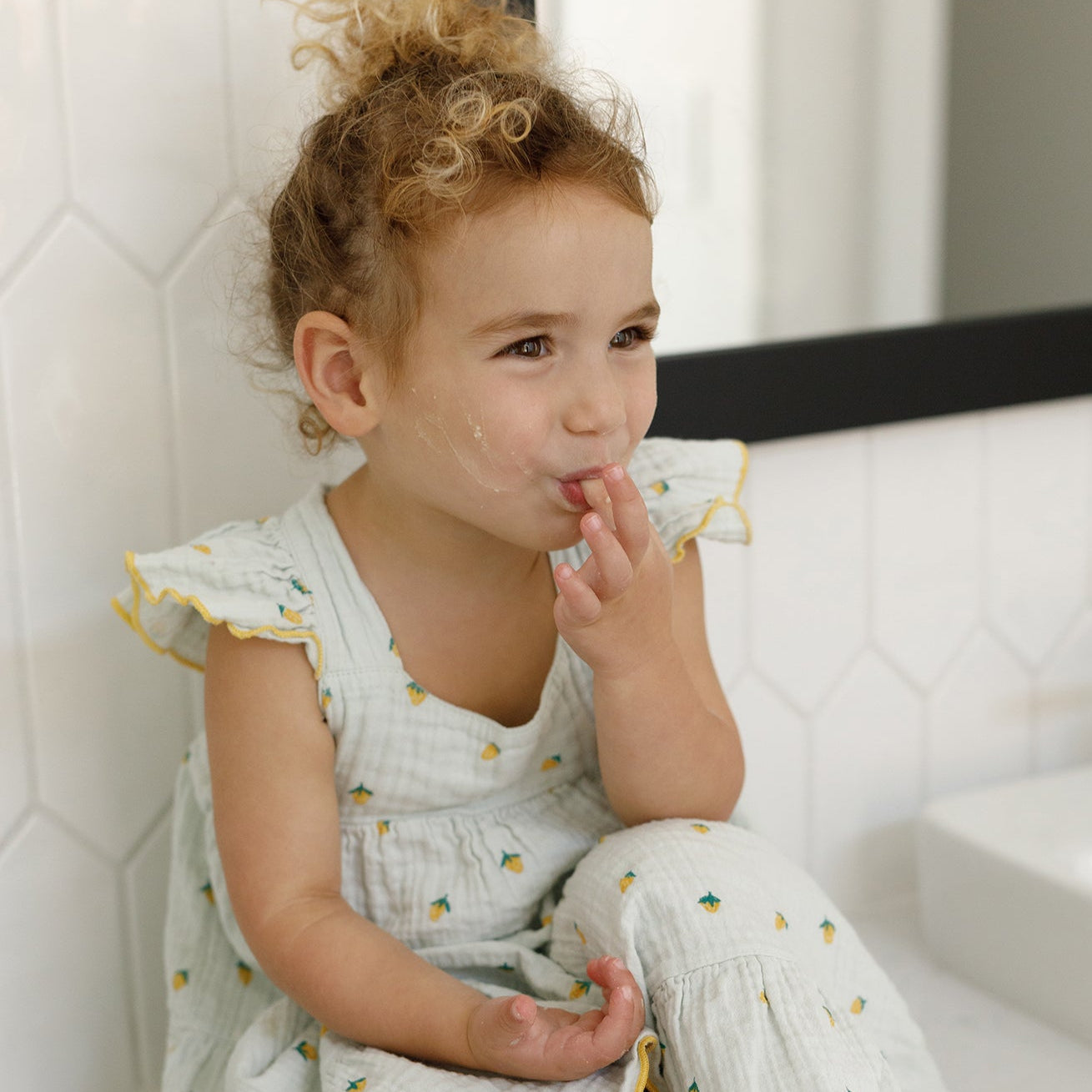 A young child with curly hair in a light dress sits in a bathroom, smiling with food on their cheek—a perfect moment for gentle cleansing using Wild Mamas’ Honey Bee (Raw Honey)-Wild Bison Tallow Balm. White tiles and a sink are visible behind.