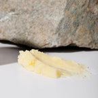 A smear of pale yellow Wild Mamas Honey Bee (Raw Honey)- Wild Bison Tallow Balm is spread on a white surface, with a large, rough-textured stone in the background.
