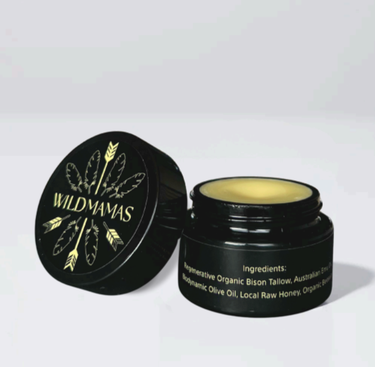 A black jar of Wild Mamas Honey Bee (Raw Honey)–Wild Bison Tallow Balm, with a feather- and arrow-decorated lid, is shown open to reveal yellow balm inside. Ingredients like wild bison tallow appear in white text on the jar.