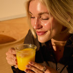 A woman with blonde hair smiles with her eyes closed, holding a clear glass cup of The Fullest Saffron Latte 30-Day Supply. Wearing a dark shirt and gold rings in warm sunlight, she savors this caffeine-free drink for mood support.
